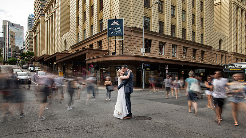 Bride and Groom stop in the middle of a city road while people rush past.