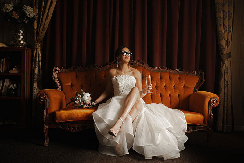 Photo by Tom Judson Photography of Bride with sunglasses and champagne in hand sits on ornate red sofa.