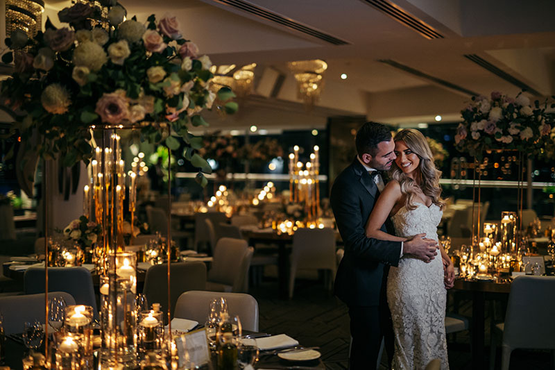 Happy Bride and Groom hold eachother in an opulent function room filled with flowers and tea lights.