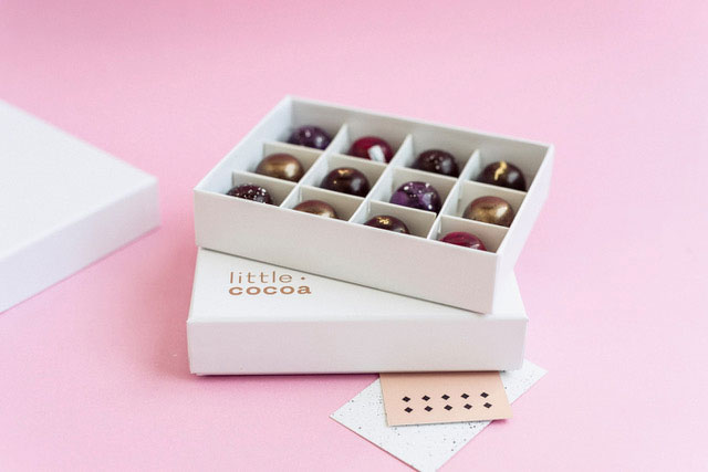 Twelve exquisite handmade chocolates in a white gift box with the name Little Cocoa on the box, on a pink background