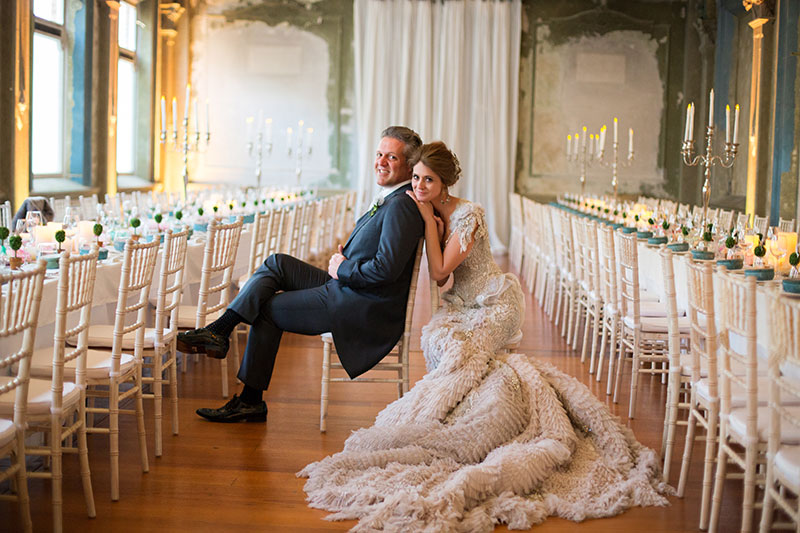 Bride in extravagent wedding gown poses with Groom inside an exquisite reception room.
