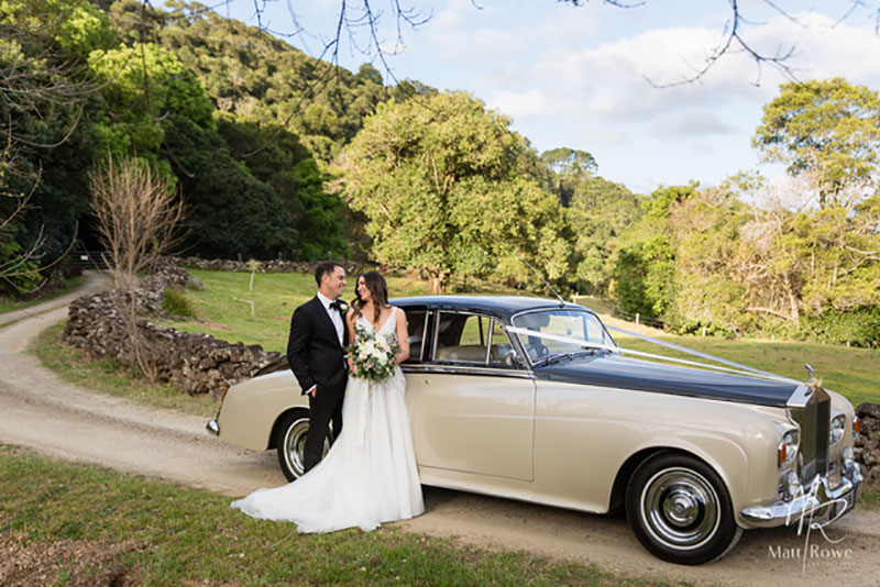 Bride and Groom pose in front of a Champagne and Black Bentley in a countryside setting.