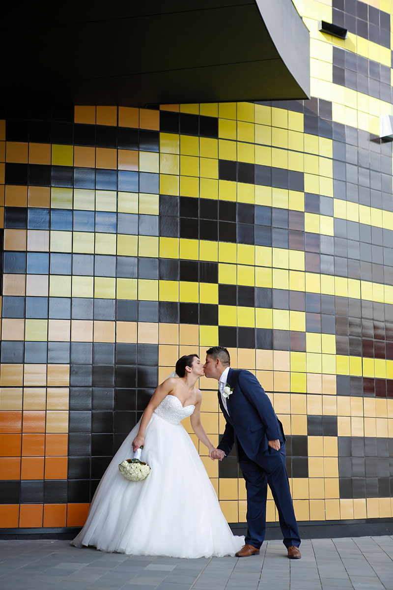 Bride and Groom kiss in front of a colourful tiled wall.