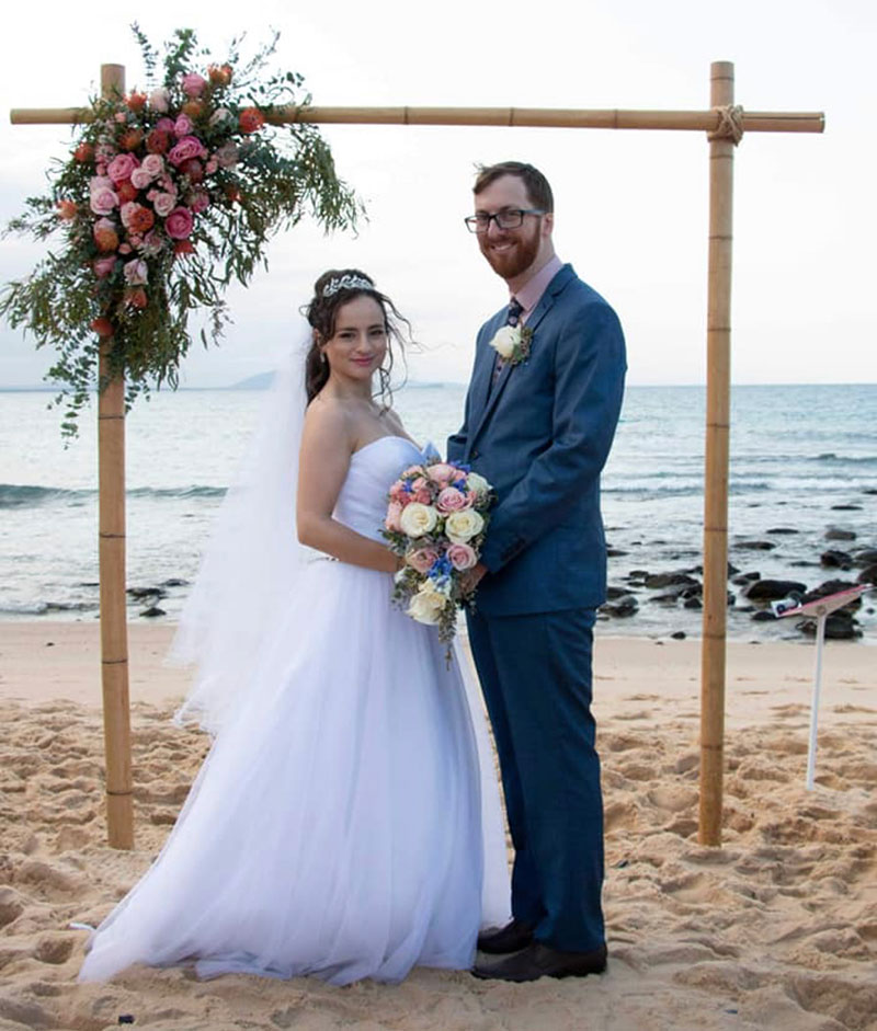 Happy Bride and Groom stand in front of their arbour on the beach.