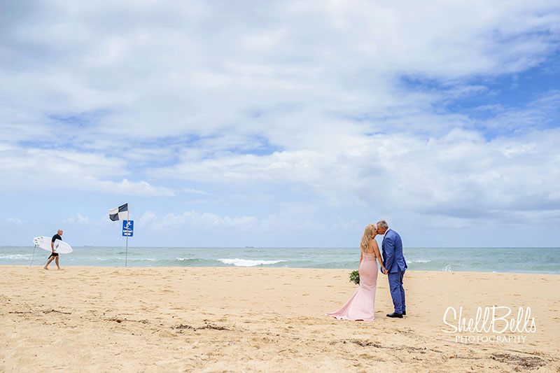 Couple kissing on beach with surfer walking in the background.