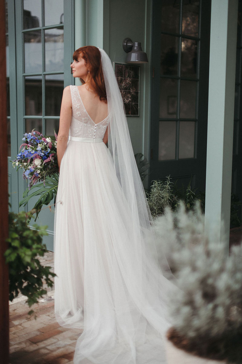 Rear view of a bride wearing a gown from French by Wendy Makin and holding flowers.