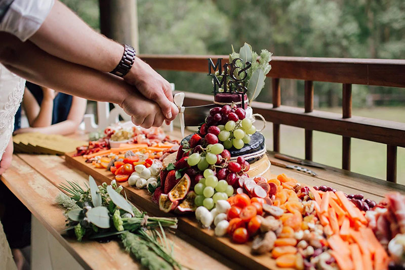 Vibrant, fresh looking wedding grazing board showing bride and groom cutting the cake made of cheese.