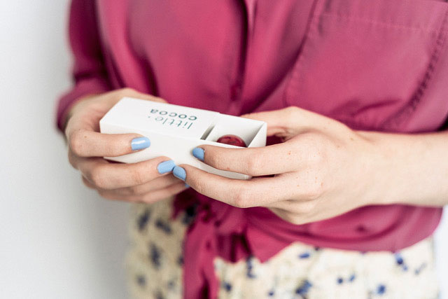 Woman with blue nailpolish and wearing a red shirt holding chocoates in a white box named little cocoa.