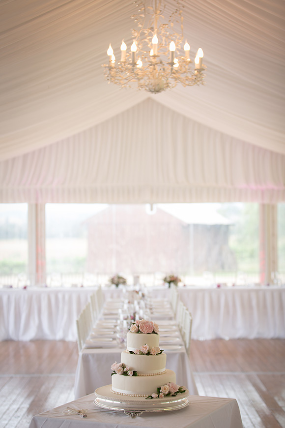 White three tiered wedding cake with pink flowers displayed under a chandelier in a room set in white.