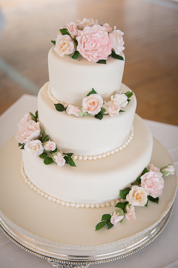 Close up of a white three tiered wedding cake with pink flowers and pearls.