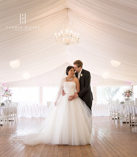 Margaret, wearing her Darb Bridal Couture gown and Michael, wearing a Black Jacket Suiting suit, together under a chandelier.