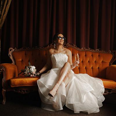 Photo by Tom Judson Photography of Bride with sunglasses and champagne in hand sits on ornate red sofa.