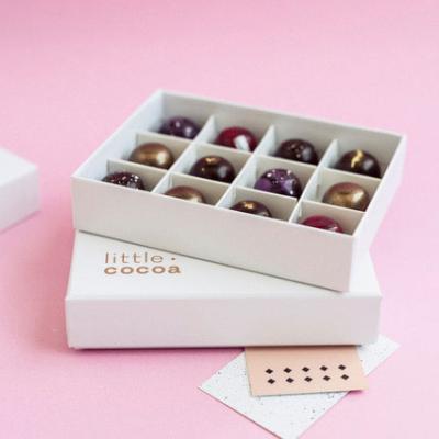 Twelve exquisite handmade chocolates in a white gift box with the name Little Cocoa on the box, on a pink background