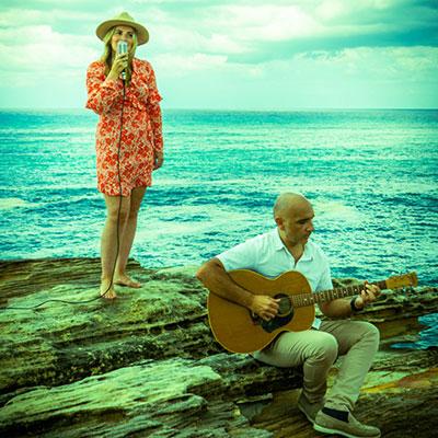 Hayley singing and Cesar playing the guitar on a rock overlooking the ocean.